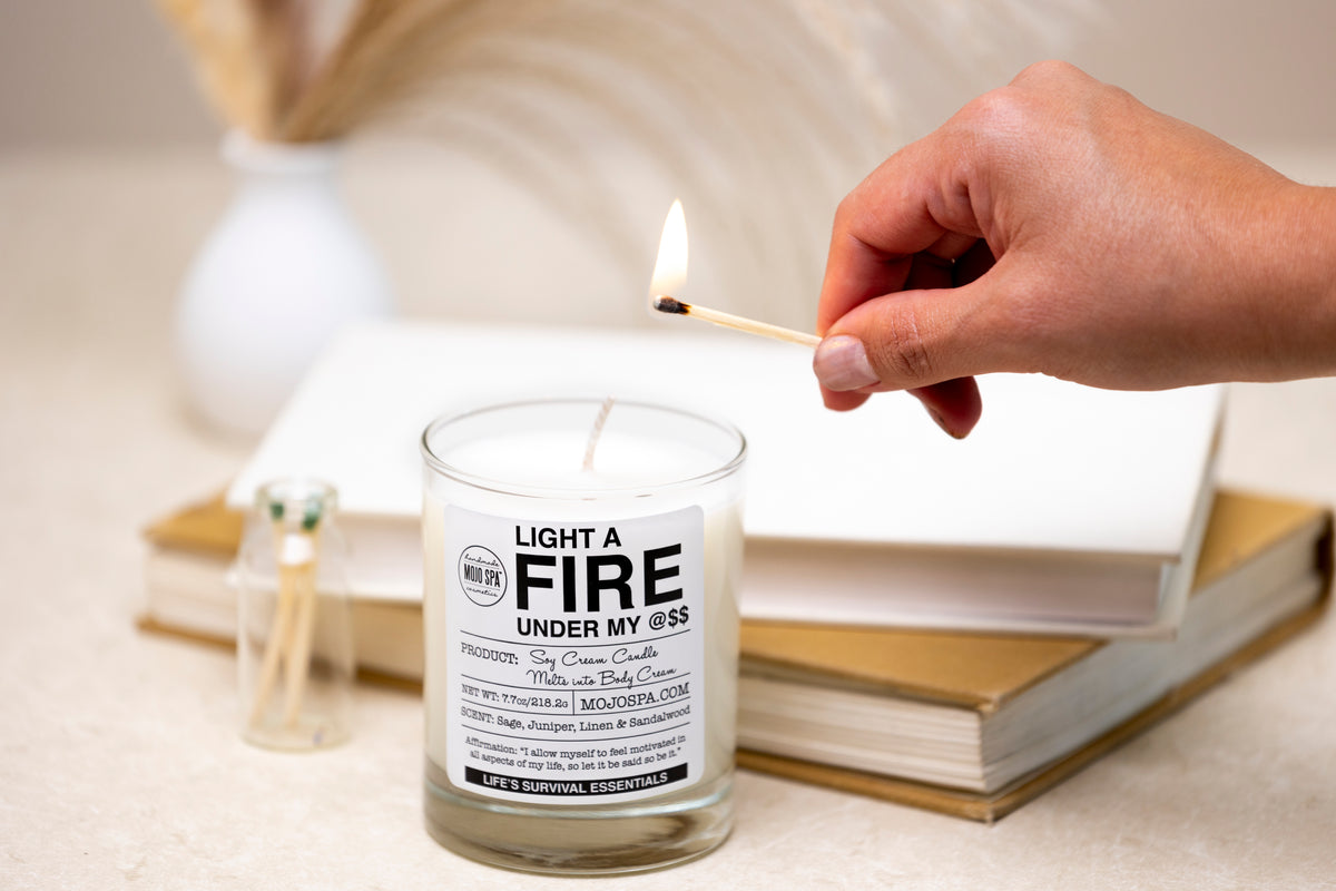 A hand holding a lit match to the wick, with books and a jar of matches behind