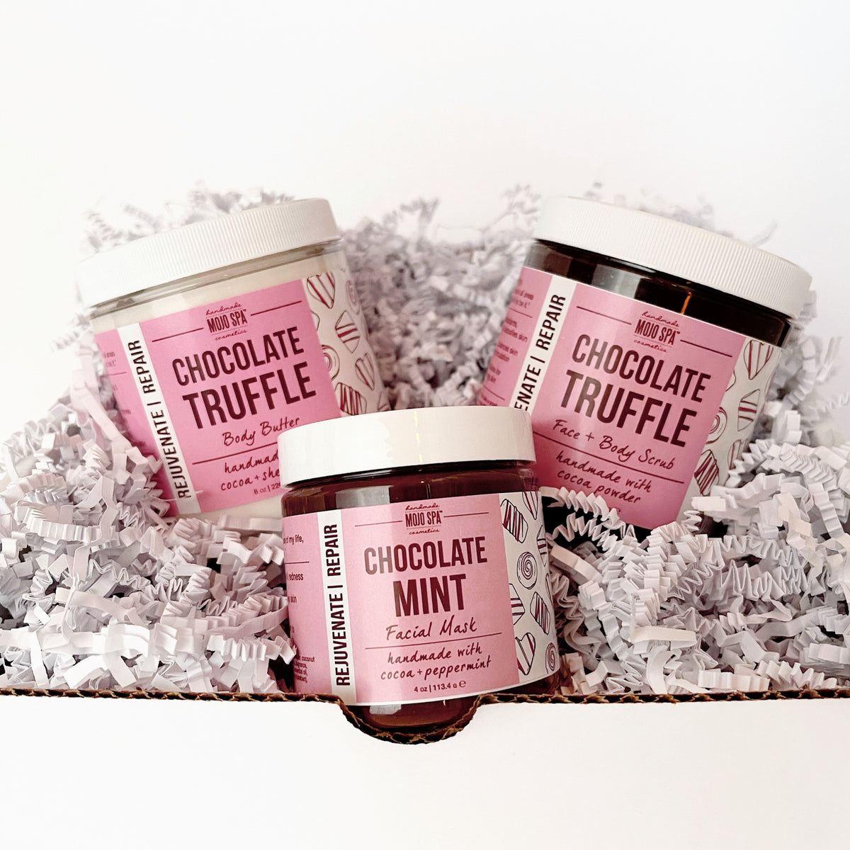 The three jars nestled in white paper shred in an open box, with the facial mask jar at the front