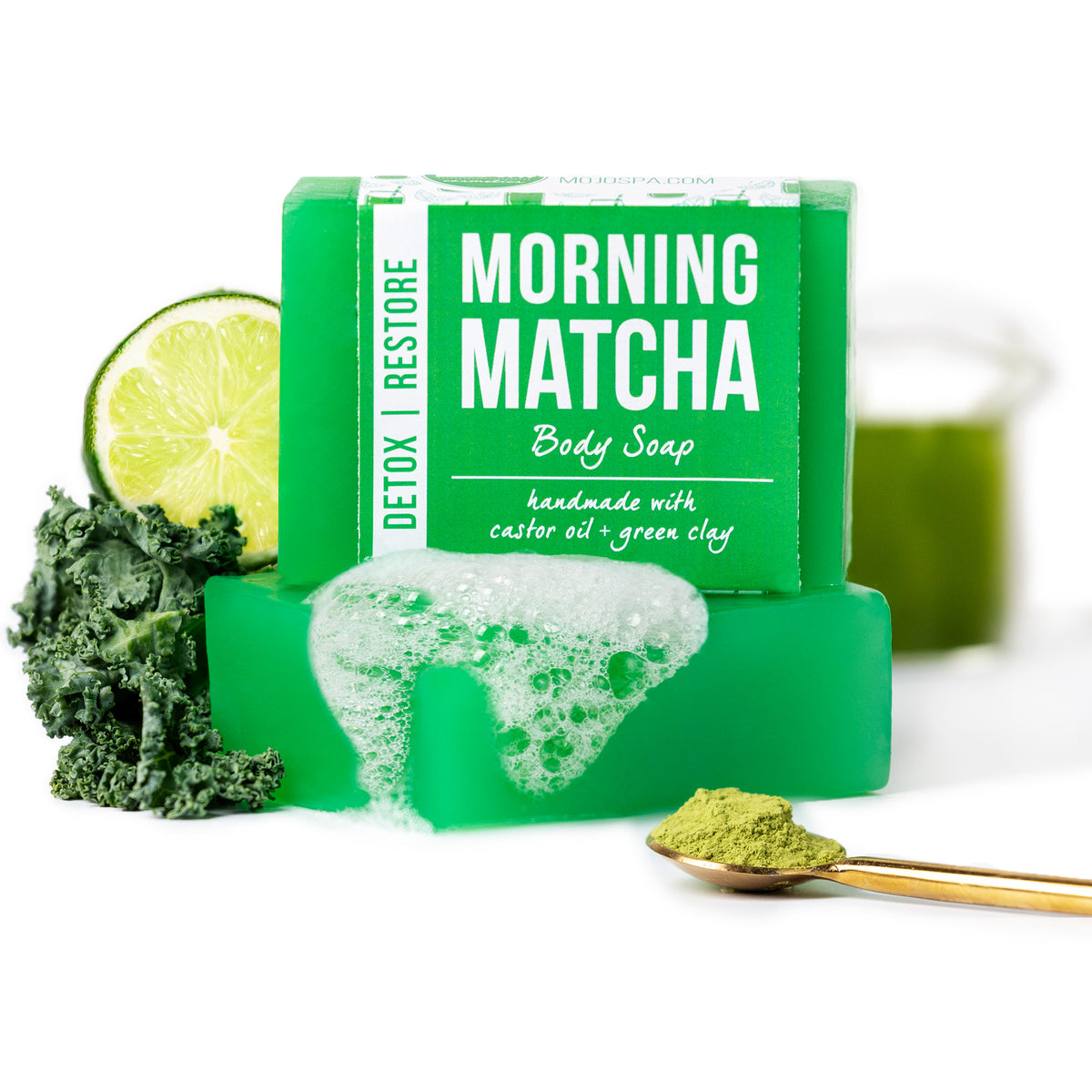 The soap bar stacked on a foam-covered bar, with a lime half, kale, a spoon of matcha powder and a glass of green liquid behind