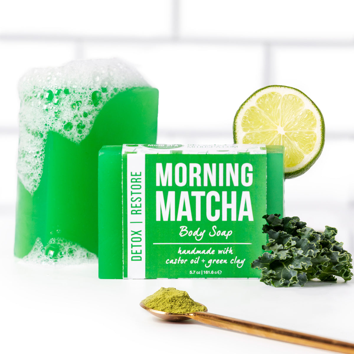 The soap bar with a foam-topped bar behind, beside a lime half, kale and a spoon of matcha powder