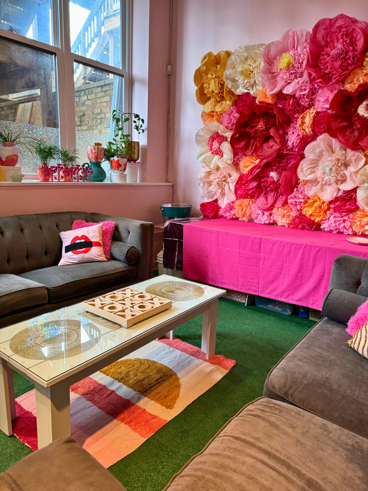 A corner of the party lounge with a couch, a pink table and a wall of paper flowers