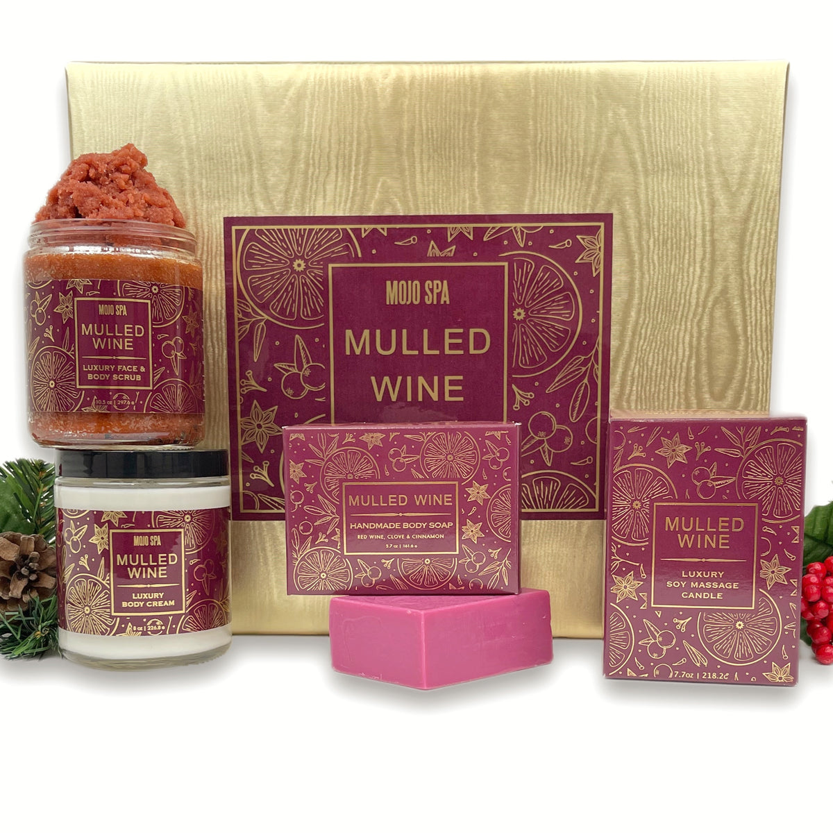 The set contents in front of the gold box, a scrub jar, a body cream jar, a soap bar with its carton and a candle box, with pine and red berries at the sides