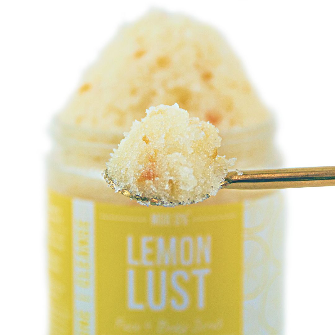 A gold spoon lifting a scoop of scrub in front of the jar