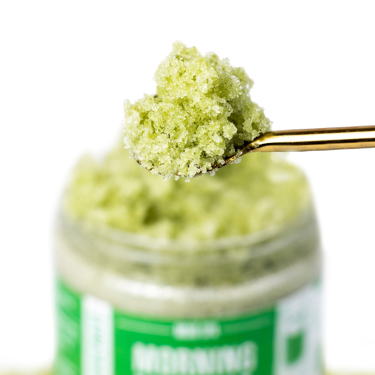 A close-up of a gold spoon lifting a scoop of green scrub above the jar