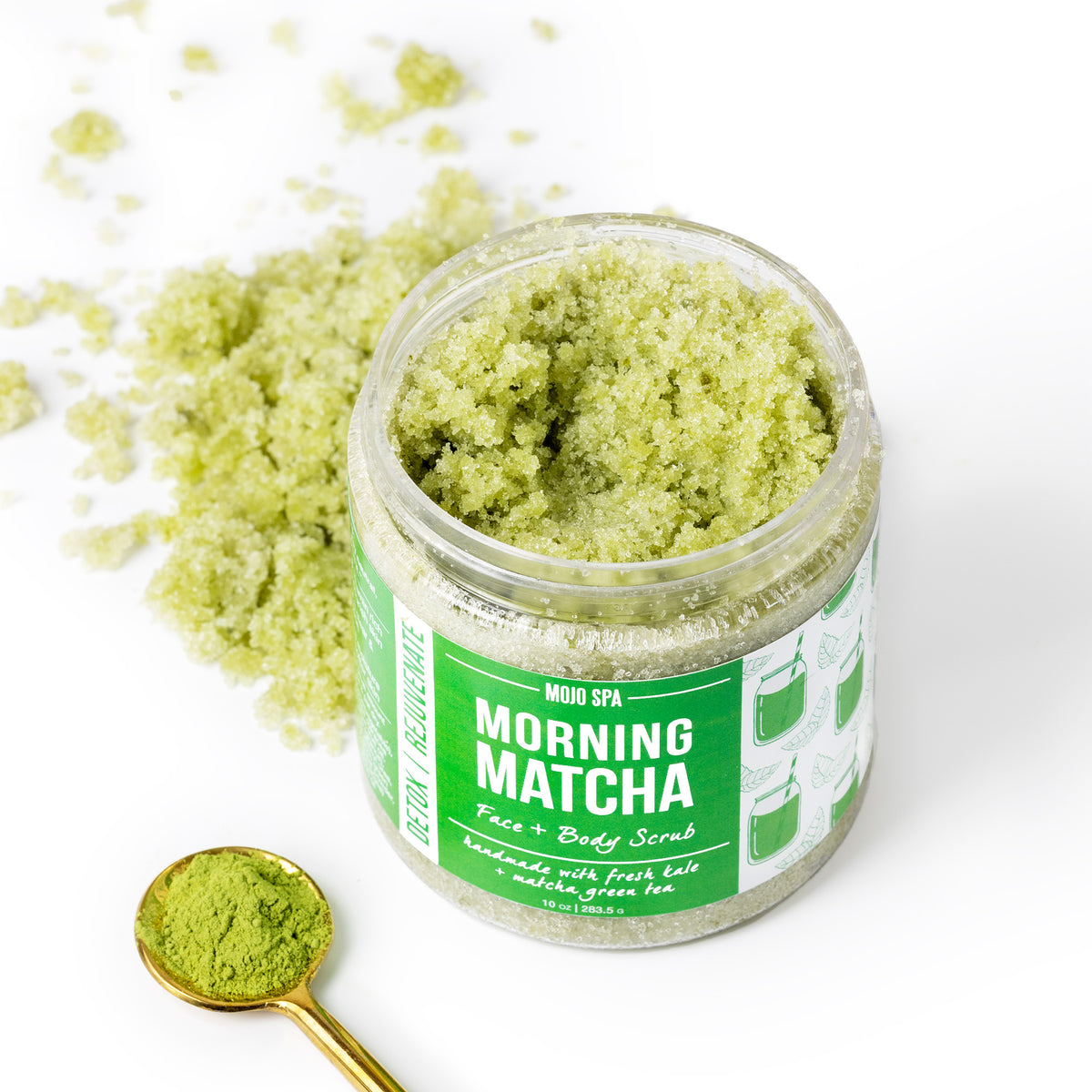 The open jar seen from above, with a spoon of matcha powder and loose scrub scattered beside it